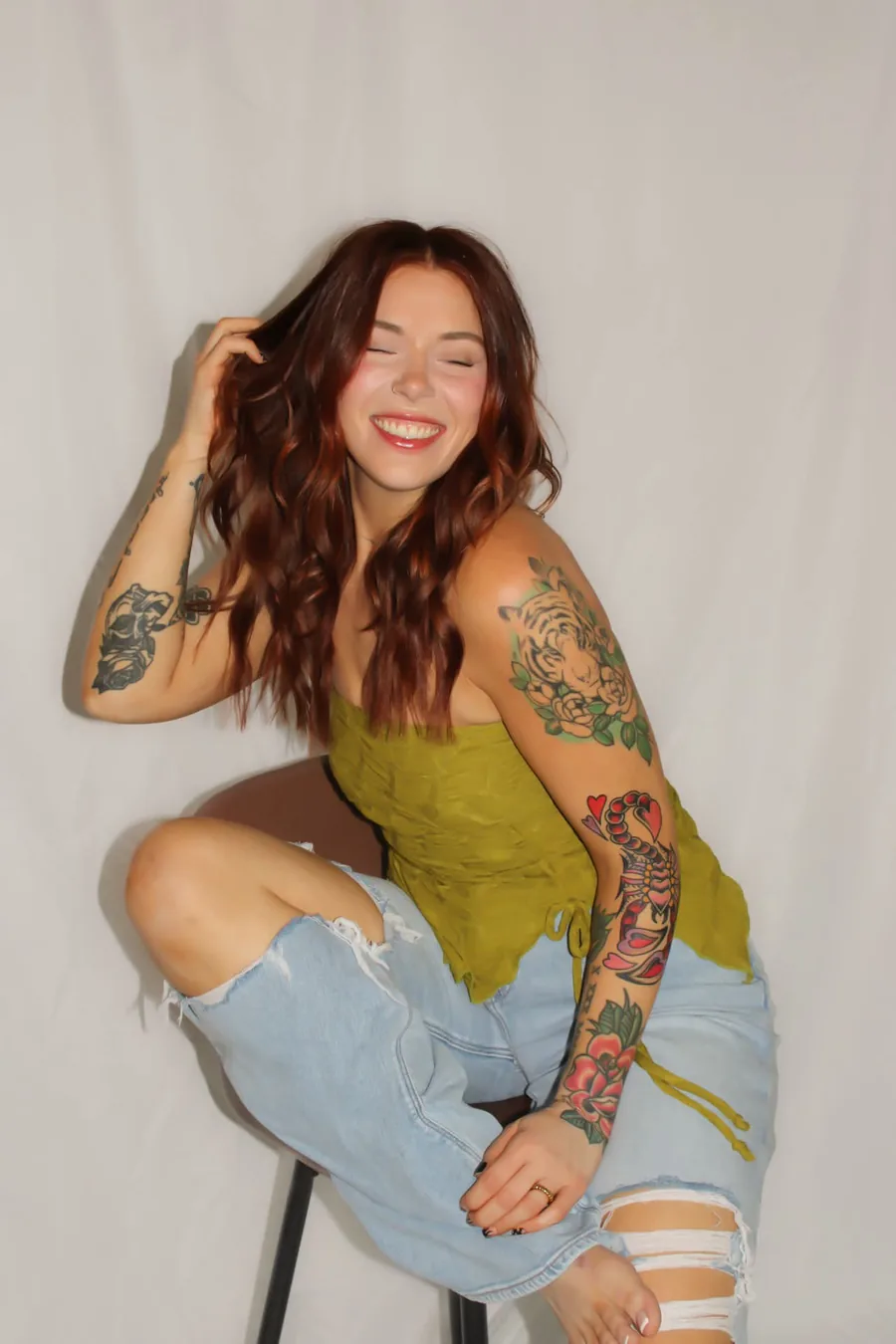 Kate McMillen laughing on a stool in a green top and light denim, her auburn waves loose over one shoulder.